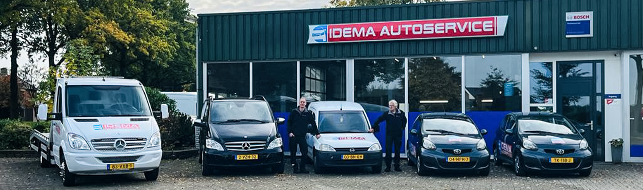 Het pand van Idema Autoservice in Emmen met twee mannen tussen het wagenpark: een lierwagen, een bestelbus en drie personenauto's